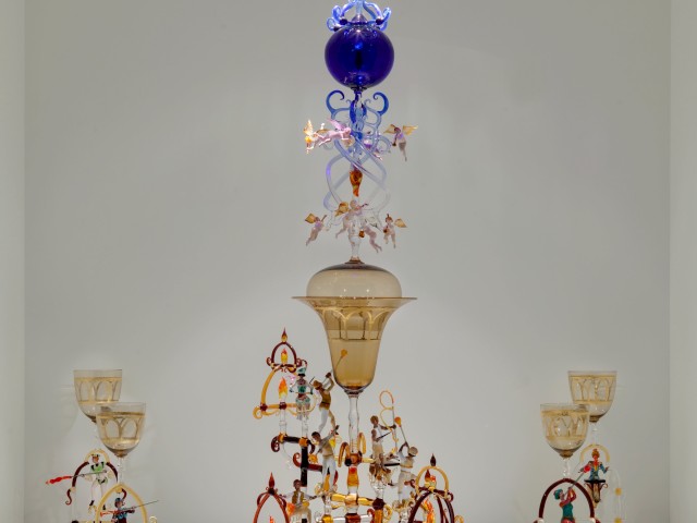 An intricate glass sculpture with colorful ornate elements and multiple tiers, displayed against a plain background.