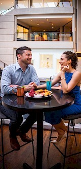 A man and woman are sitting at a table in a modern setting, enjoying drinks and a meal, and engaging in conversation.