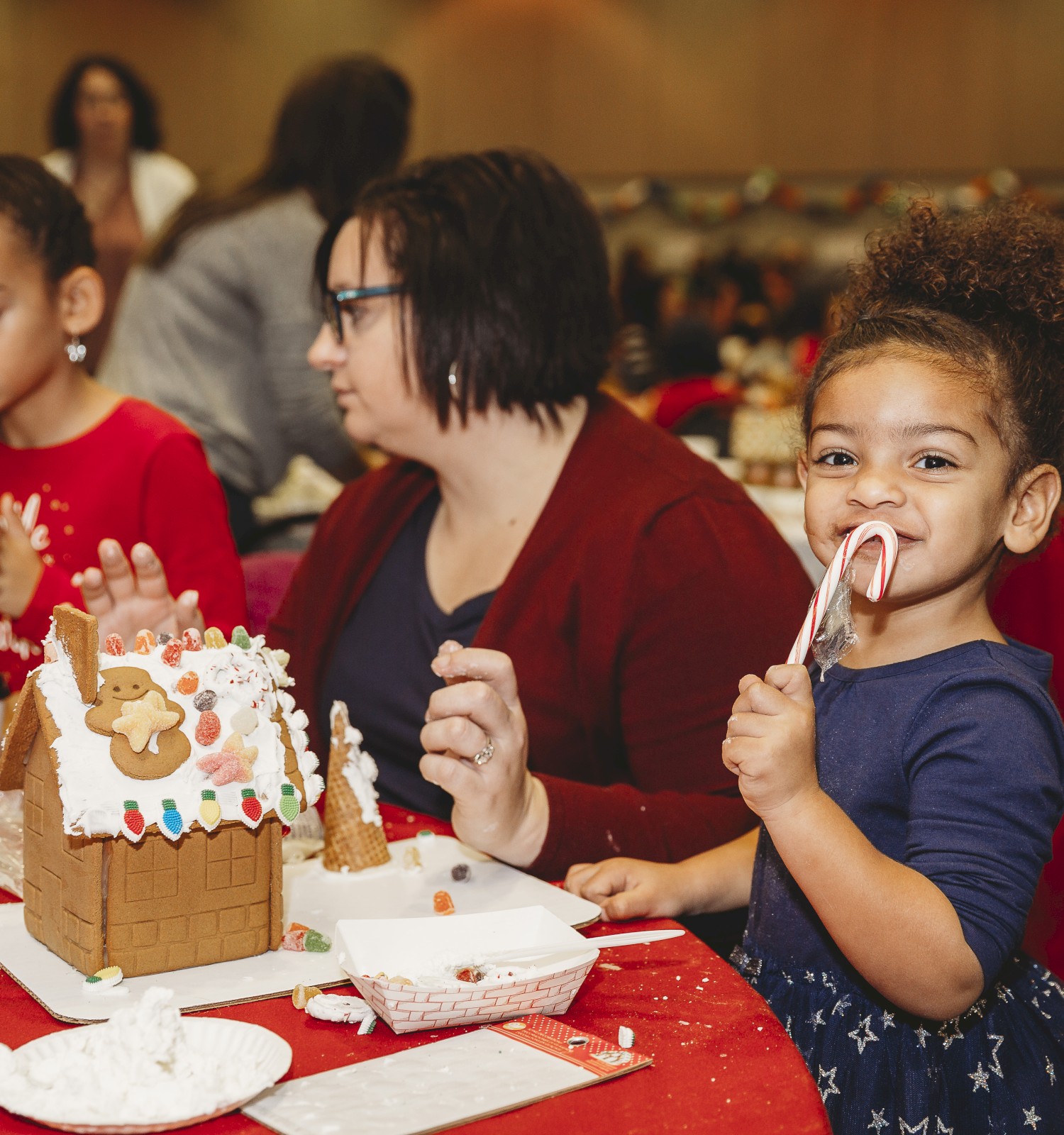 Kids at a birthday party decorating a gingerbread house, with plates, drinks, and a smiling little girl licking a spoon, festive and cheerful.