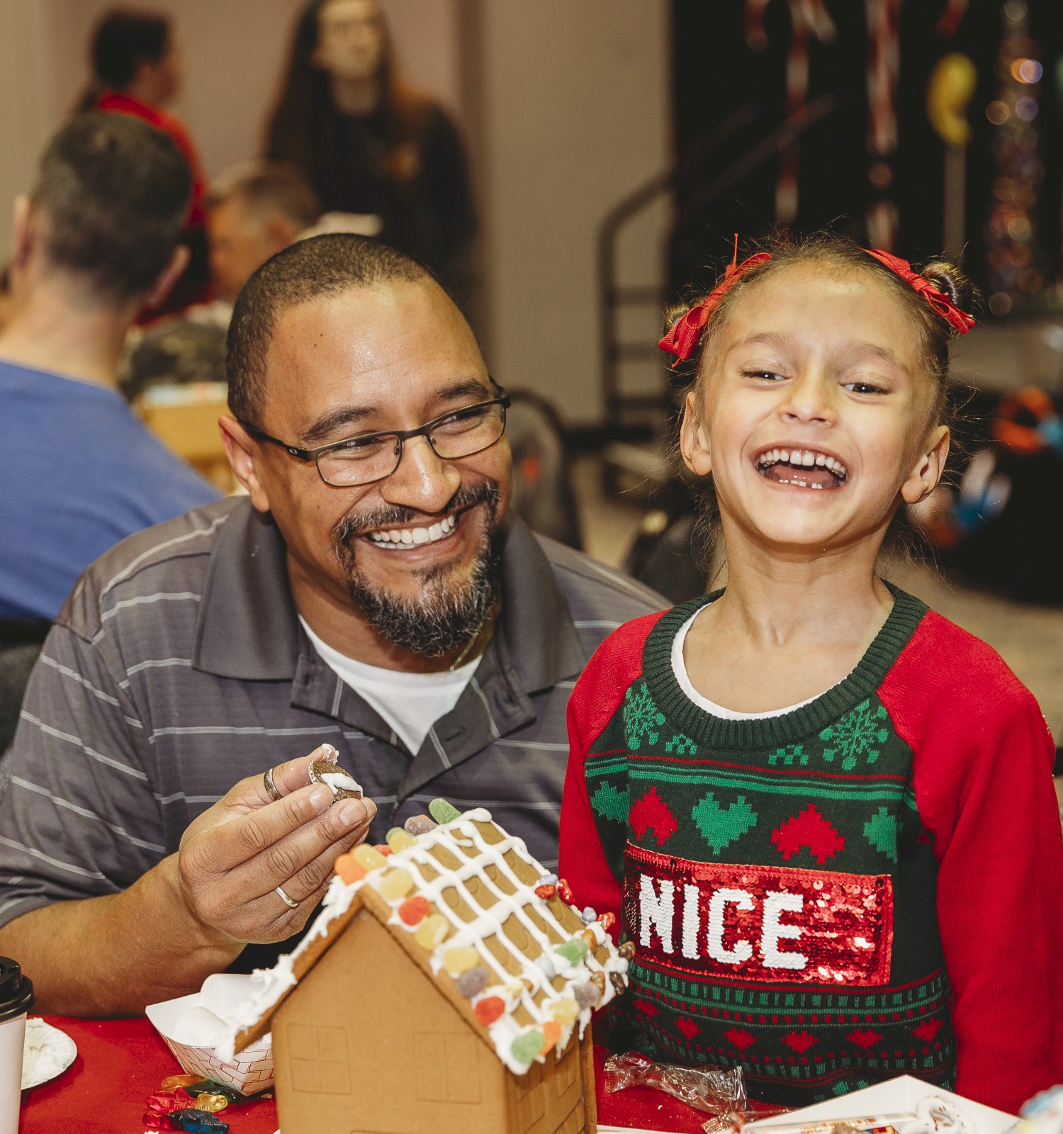 A man and a girl smile while decorating a gingerbread house at a festive table, enjoying a holiday crafting moment together.