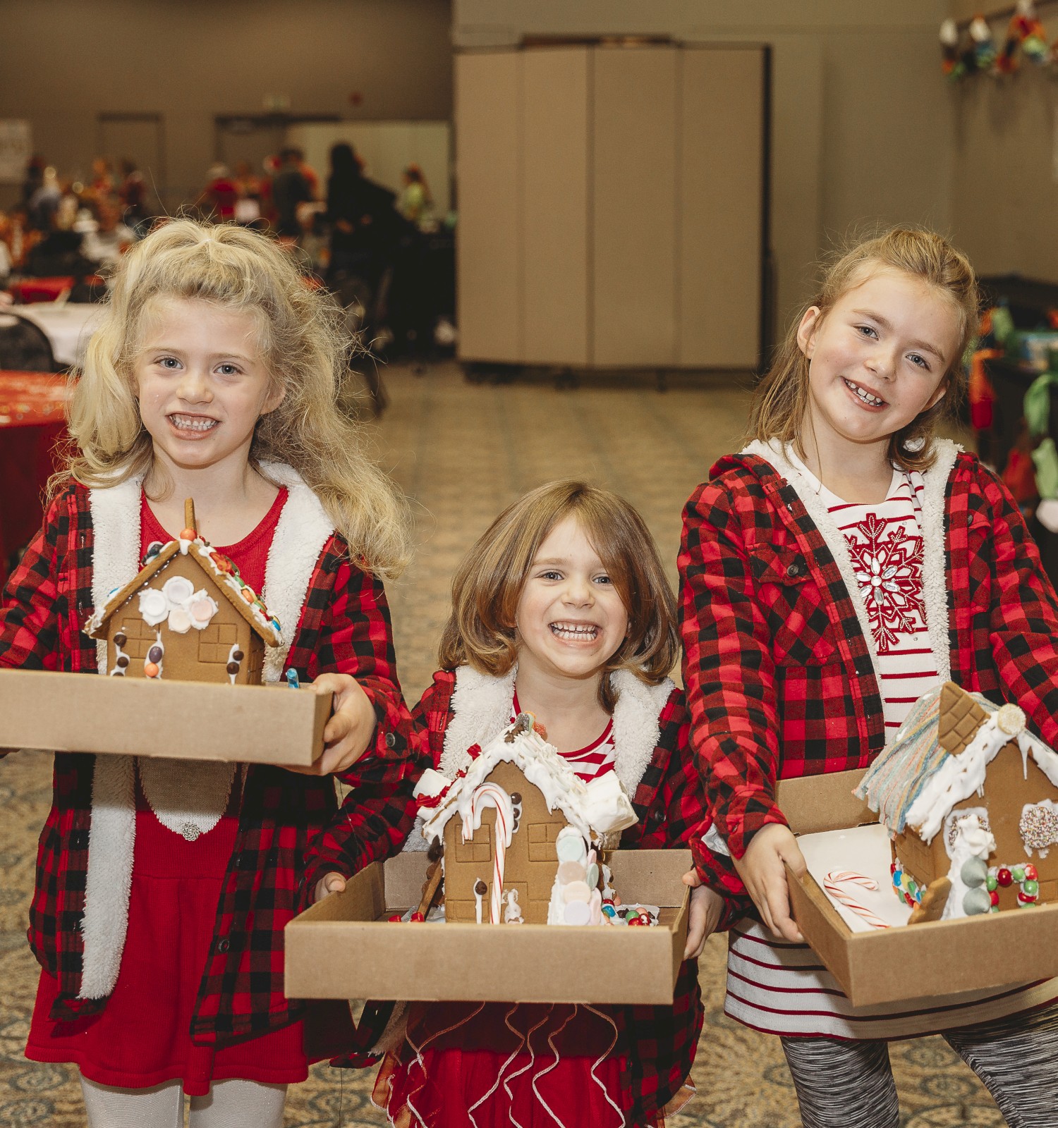 Three cheerful girls in red plaid outfits hold cardboard trays of gingerbread houses at a festive market, smiling brightly at the camera.