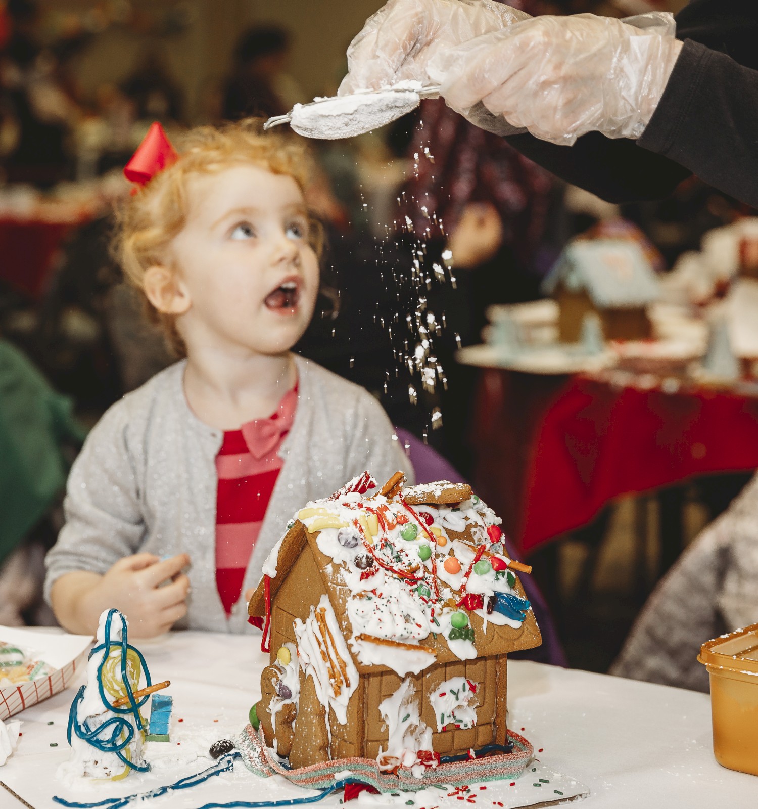 A young girl watches as a person sprinkles icing on a gingerbread house at a birthday party, with colorful decorations around.