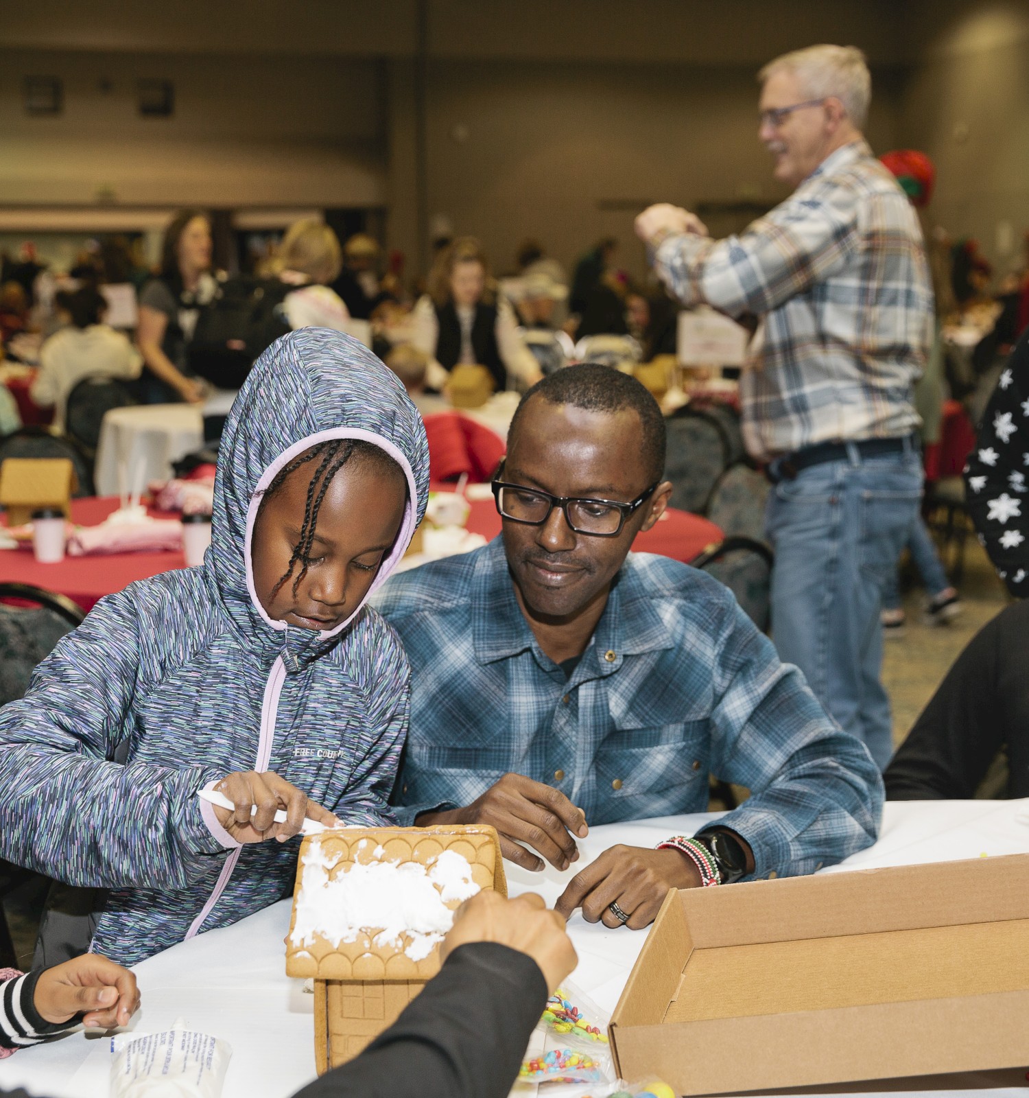 A group of people, including kids, are crafting or wrapping gifts at a table in a busy indoor event with festive vibes.