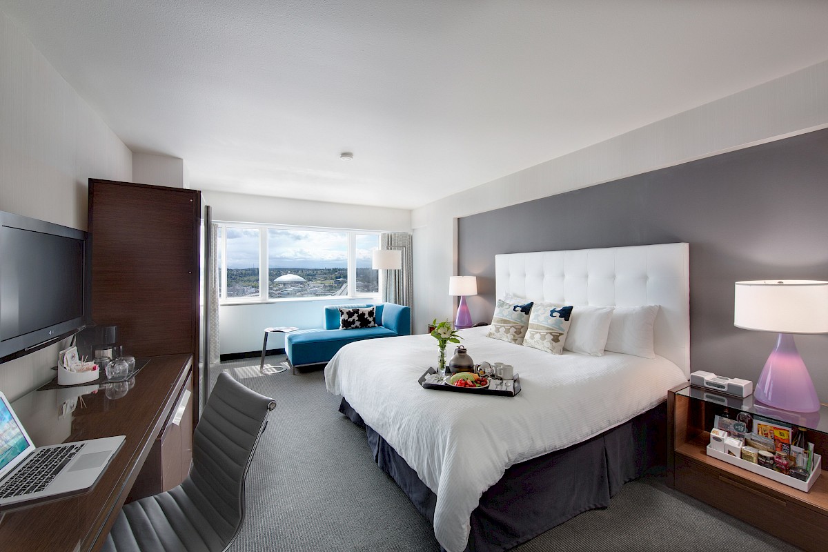 A modern hotel room with a king-sized bed, desk with laptop, TV, seating area, large window, and a tray with food and drinks on the bed.