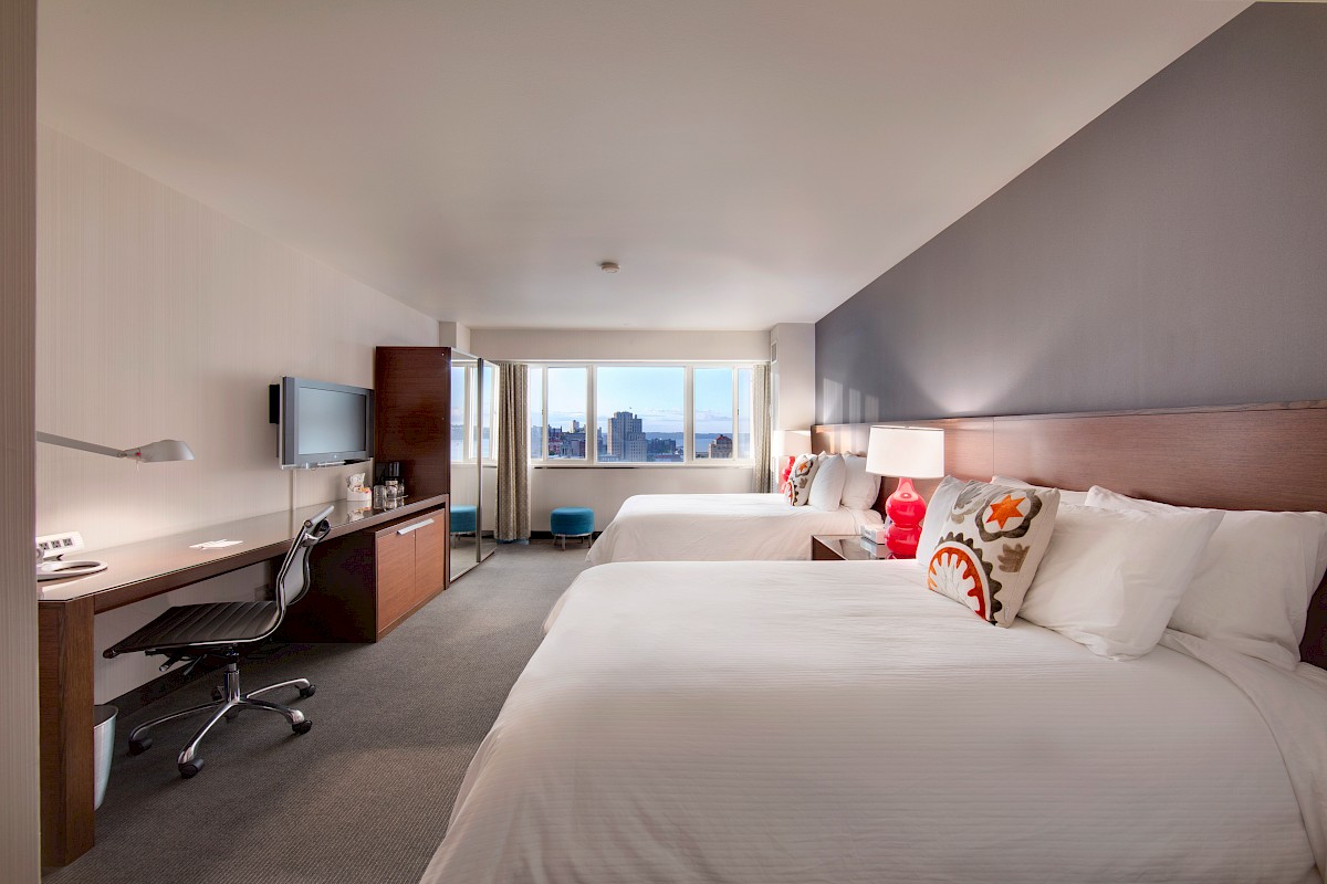 The image shows a hotel room with two beds, a desk, a chair, a wall-mounted TV, and a window with a view outside.