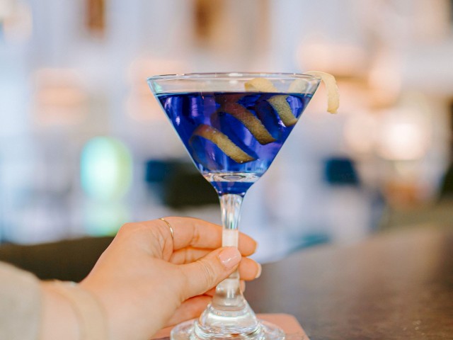 A hand holds a blue cocktail in a martini glass with lemon twists, set on a coaster in a possibly upscale bar environment.