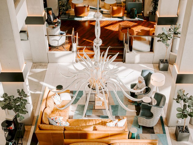 This image shows an elegant hotel lobby with modern decor, including stylish sofas, plants, a grand chandelier, and a checkered floor pattern.