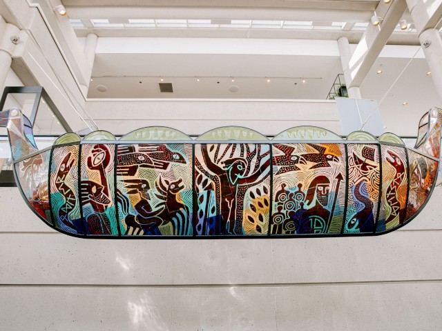 A colorful, stained glass artwork depicting various figures and patterns, displayed inside a modern, white-walled building with high ceilings.