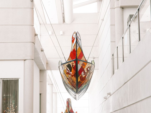 The image shows three colorful, boat-like sculptures suspended from the ceiling in a modern, white indoor space with skylights.
