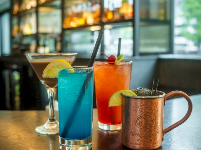 A variety of colorful cocktails, including a martini, blue drink, red drink, and a copper mug, displayed on a bar counter in a modern bar setting.