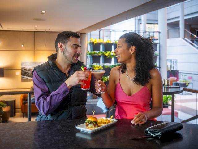 Two people are sitting at a bar, toasting with drinks. There's a plate with a burger and fries in front of them, and they appear to be smiling.