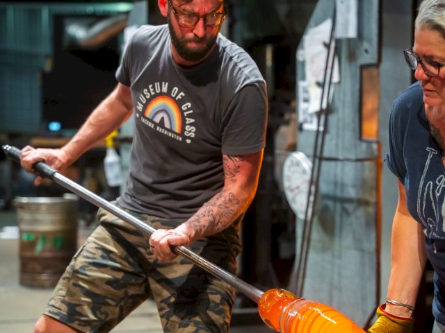 Two people are working in a glassblowing studio, shaping molten glass with a rod while wearing protective gear including gloves and glasses.