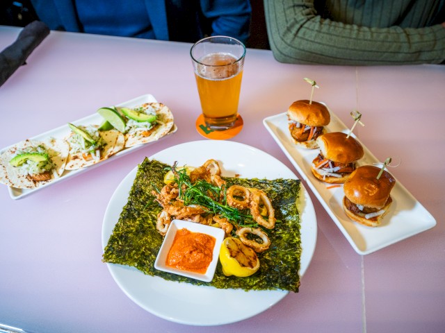 The image shows a table with various dishes: tacos, mini burgers, a calamari dish with a dip, and a glass of beer.