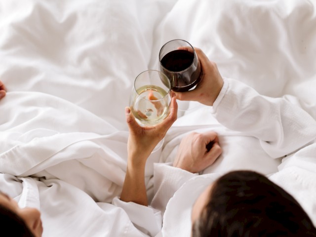 Two people are relaxing in bed, wearing white robes, and toasting with glasses of wine, one white and one red.