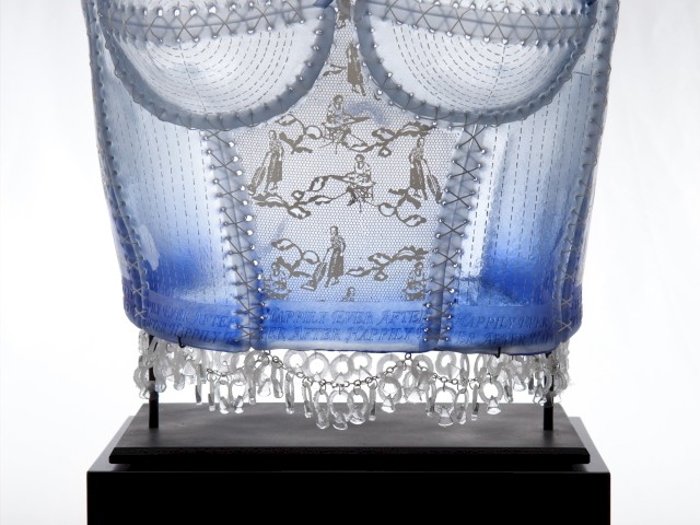 The image shows a blue and transparent sculpted corset displayed on a black pedestal, featuring intricate details and delicate craftsmanship.