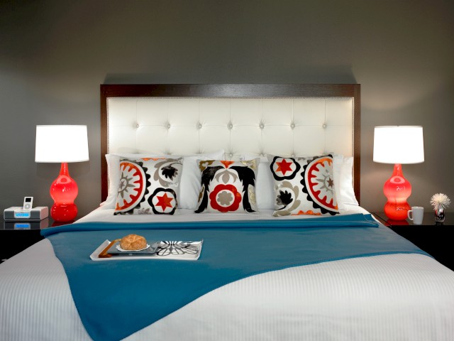 A neatly arranged bed features colorful patterned pillows, red bedside lamps, a blue throw, white sheets, a white headboard, and nightstands.