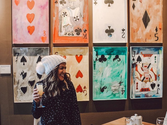 A person wearing a beanie and holding a drink sits at a table in front of a wall decorated with large playing card artworks.