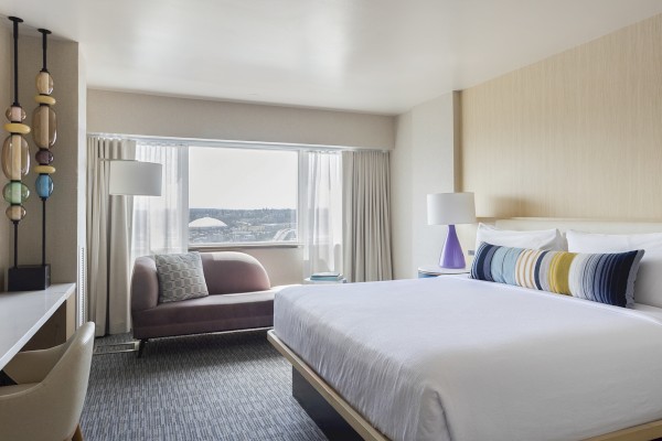 This is a bright hotel room with a large bed, colorful pillows, a window view, and modern decor including a purple lamp and a art piece.