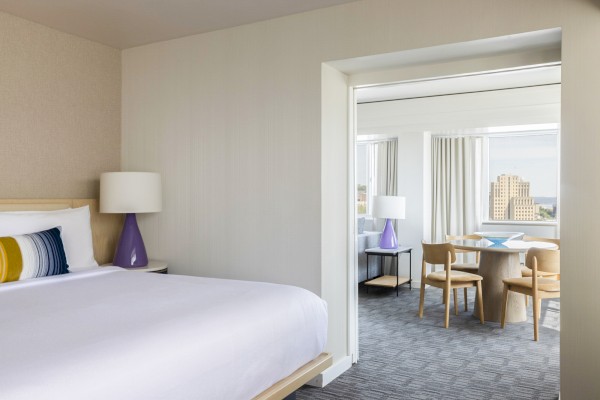 A modern hotel room with a bed, purple lamps, and a view of a cityscape from large windows.