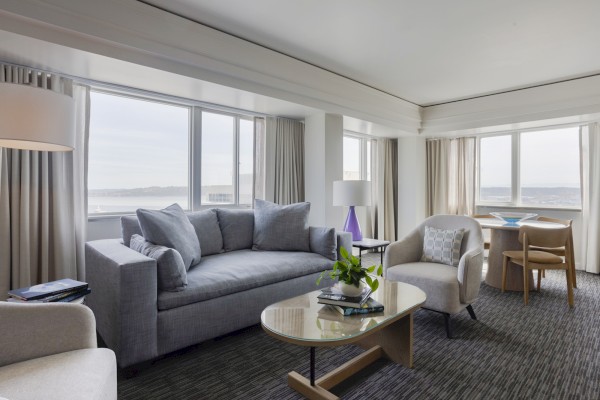 A bright, modern living room with large windows, a gray sofa, armchair, coffee table, and view of water outside.
