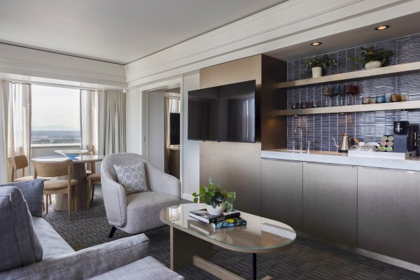 This is a modern, stylish living room and kitchenette with large windows, a TV, a dining area, and cozy seating.