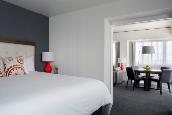 A modern hotel room with a large bed, lamps, and a view into a sitting area with a dining table and chairs, decorated with bright accents.