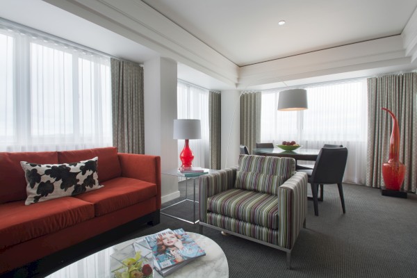 A modern living room with striped armchair, red couch, decorative lamps, and a dining area with a table and chairs by large windows.