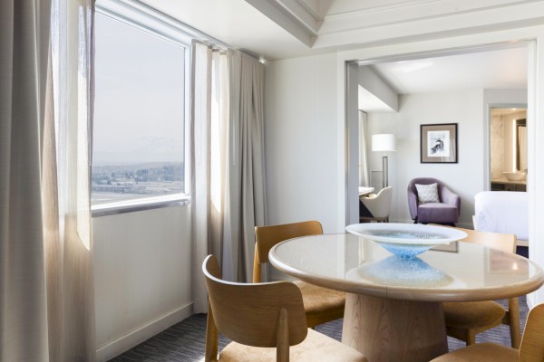 This image shows a bright, modern hotel suite with a round wooden dining table, chairs, and a view of the city through large windows.