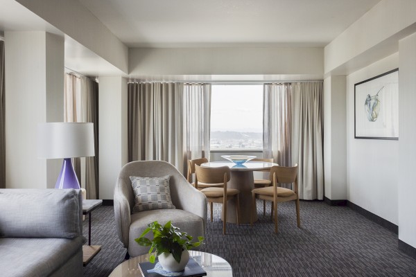 The image shows a modern, well-lit hotel living room with a round dining table, chairs, a cozy armchair, a side table, and a large window with curtains.