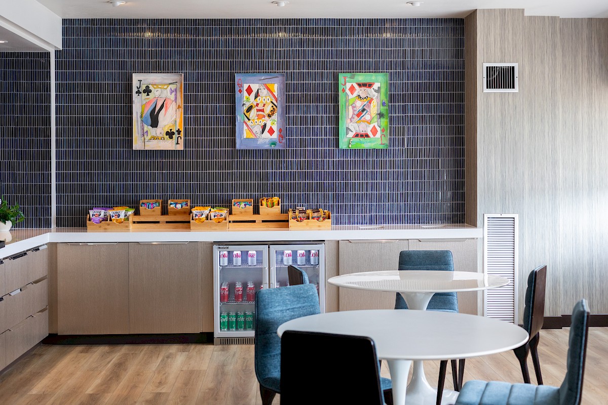 A modern refreshment area with abstract artwork, snack containers, small refrigerators, and round tables with chairs.