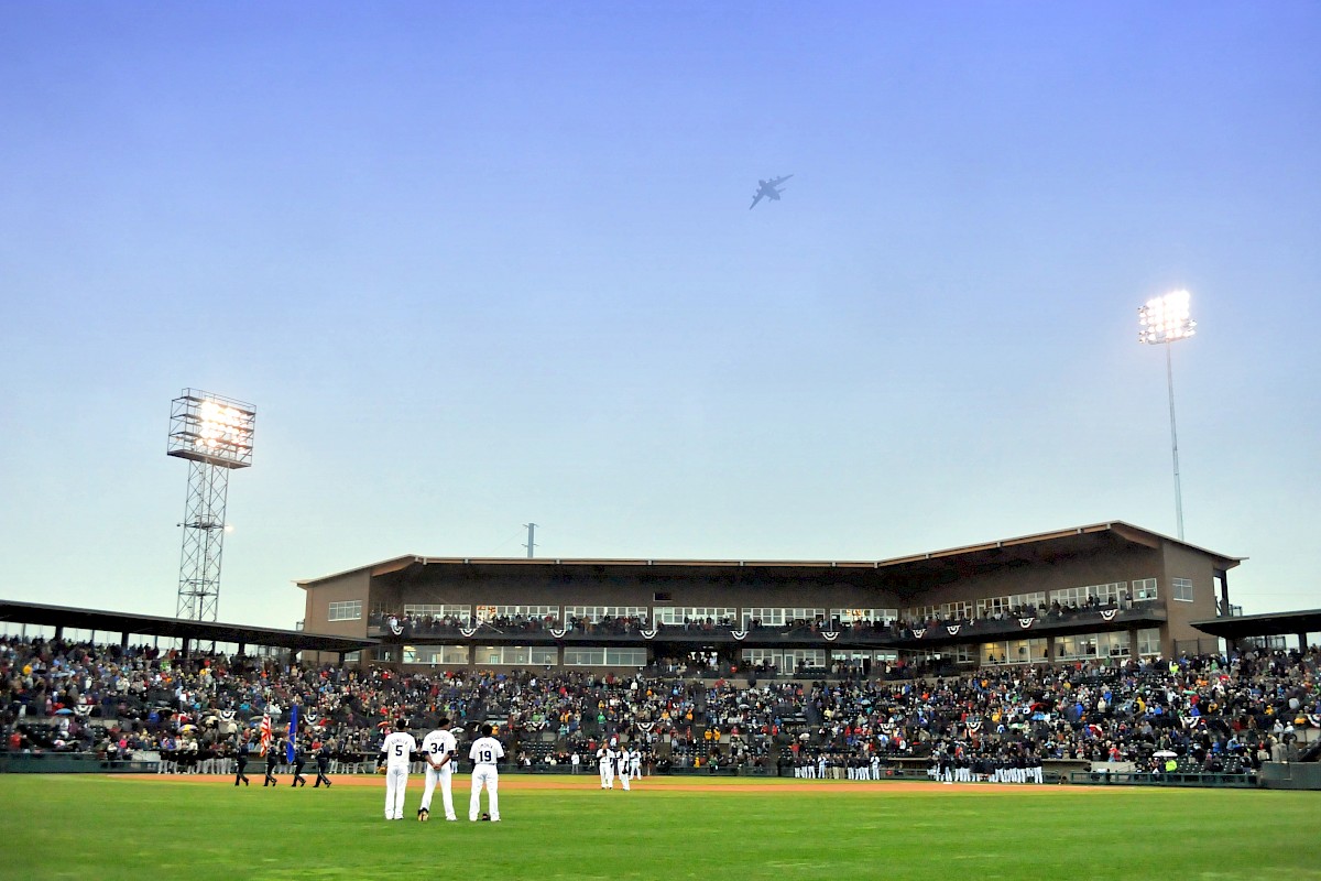 A baseball game with players on the field, a plane flying overhead, and a stadium filled with spectators under bright lights.