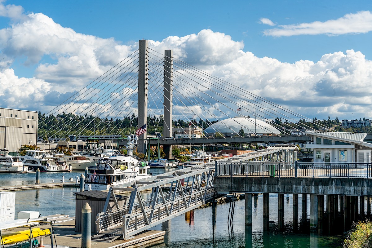 The image shows a marina with boats, a bridge, and a domed stadium in the background under a partly cloudy sky.