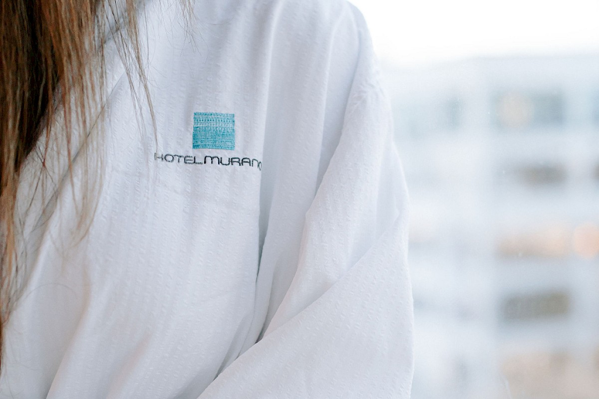 A person in a white robe is standing by a window, looking outside. The robe has a logo with text on it.