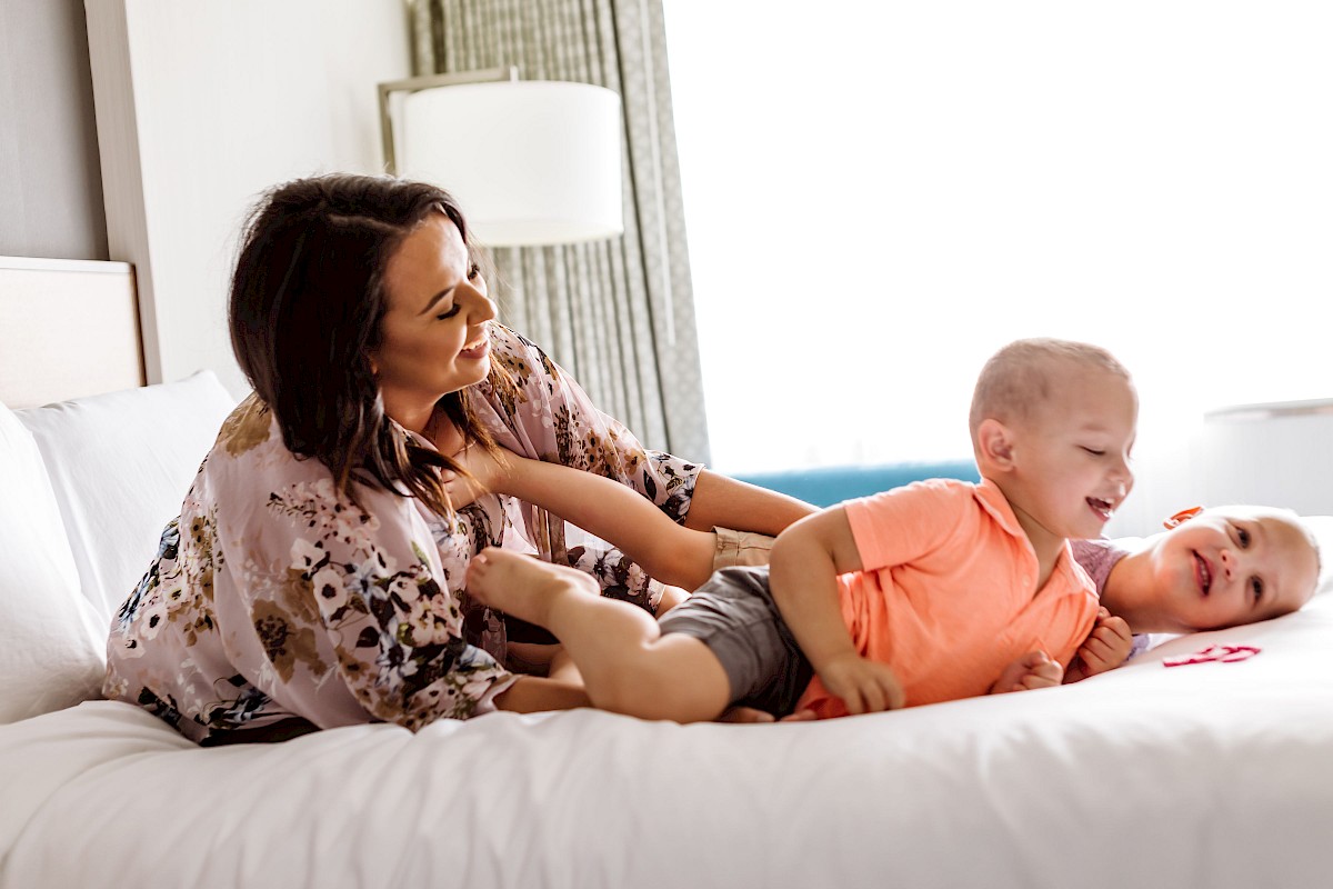 A woman wearing a floral robe playfully interacts with two laughing children on a bed in a bright room.