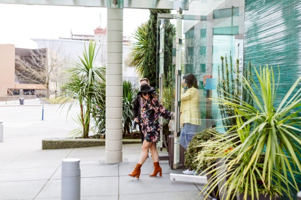 A fashionable woman in a floral dress, cowboy boots, and a hat is entering a building with two people inside.