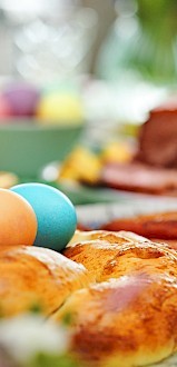 A festive table with colored eggs, braided bread, and a glass. In the background, there's a dish and more colored eggs in a bowl.