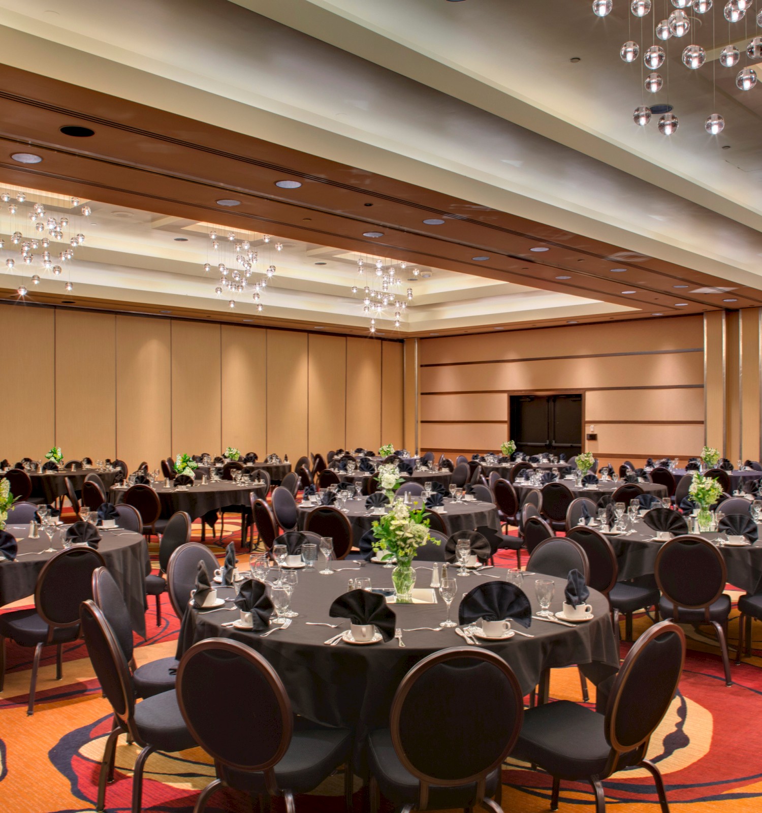 A decorated banquet hall set up for an event, featuring round tables with black tablecloths, chairs, and floral centerpieces under elegant lighting.