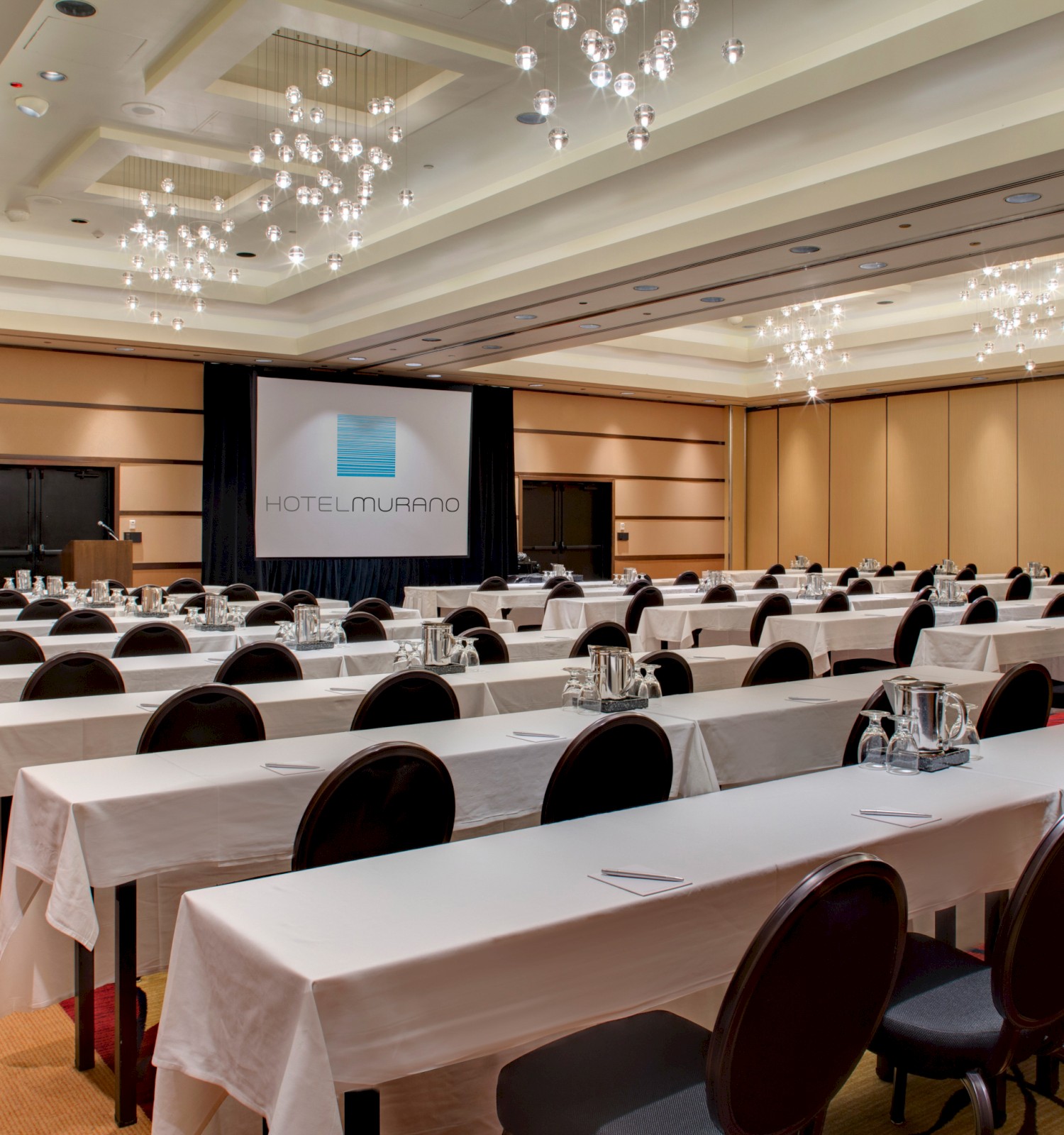 A large conference room in Hotel Murano with arranged tables and chairs, notebooks, water pitchers, and elegant ceiling lights.
