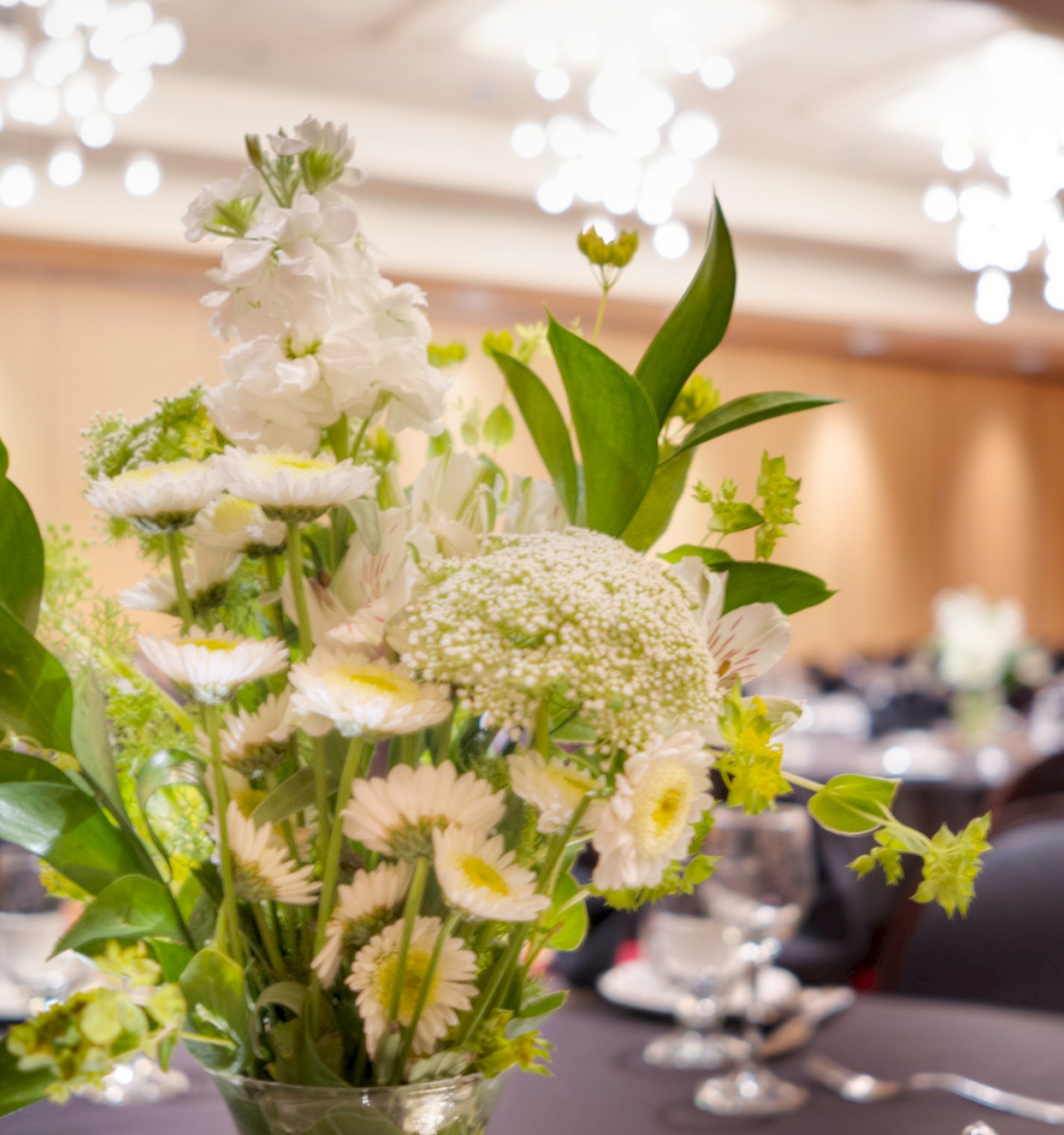 A bouquet of white and green flowers in a vase is at the center of a banquet table in a decorated venue.