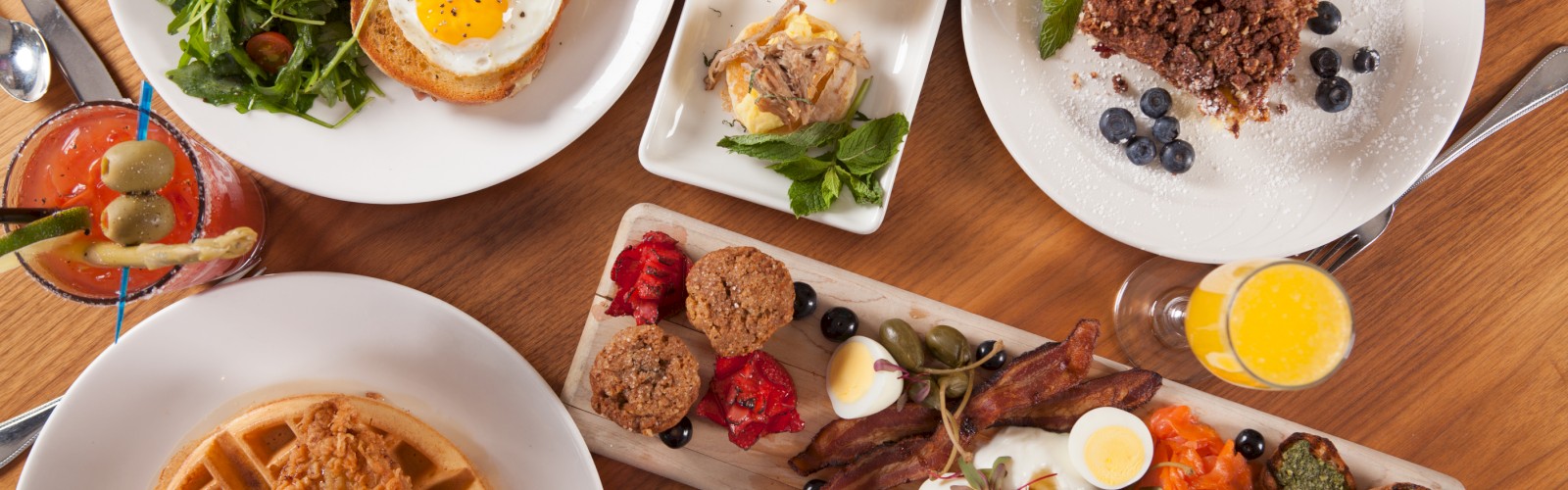 An assortment of breakfast dishes: waffles with chicken, fried egg on toast, pastries, salad, and drinks, displayed on a wooden table.