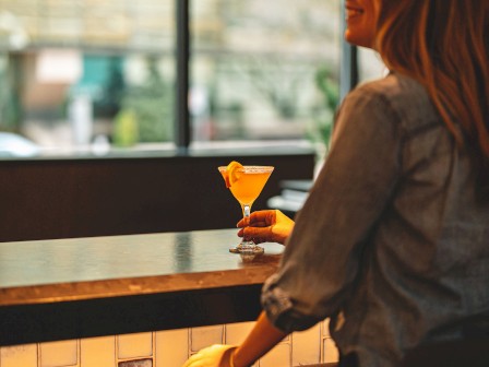 A person sits at a bar holding a cocktail, facing large windows with a blurred exterior view.