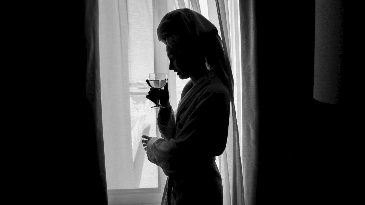 A silhouette of a person wearing a robe, standing by a window, holding a glass, with light filtering through curtains.