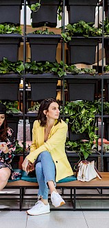 Two people sit on a bench with luggage near a vertical garden wall, in a bright interior space. One is on a phone.