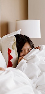 A woman peeks out from under the covers to smile at a man sleeping beside her in a cozy hotel room.