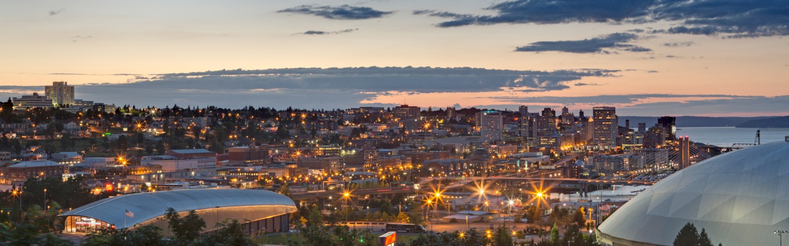A cityscape during sunset with a skyline, buildings, and a brightly lit sports dome are visible under a partly cloudy sky.