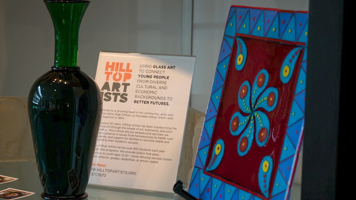 The image shows a green glass vase, a framed artwork with a blue and red pattern, and a sign for Hilltop Artists.