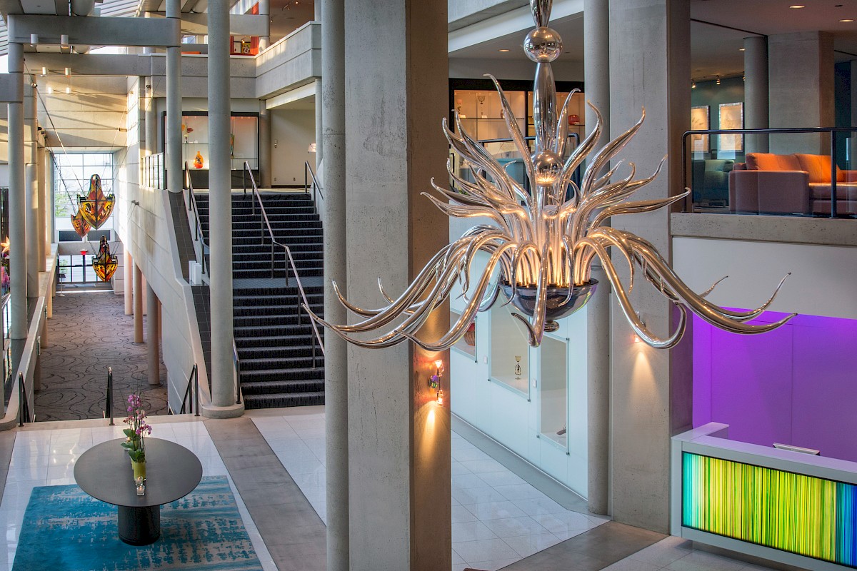 A modern lobby features a large chandelier, a staircase, vibrant lighting, and a round table with plants.