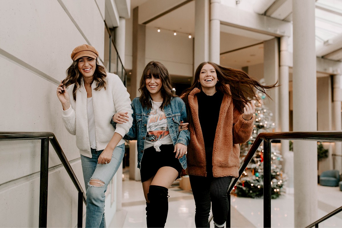 Three women smiling and walking indoors near a decorated Christmas tree, wearing winter outfits and enjoying themselves.