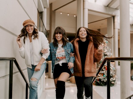 Three people are walking indoors, smiling and holding each other, with a Christmas tree in the background.