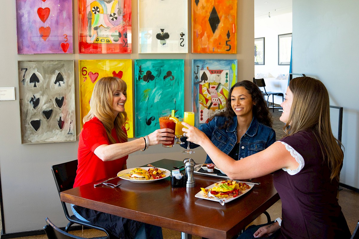 Three people toast drinks at a table with food. Large playing card art decorates the wall behind them.